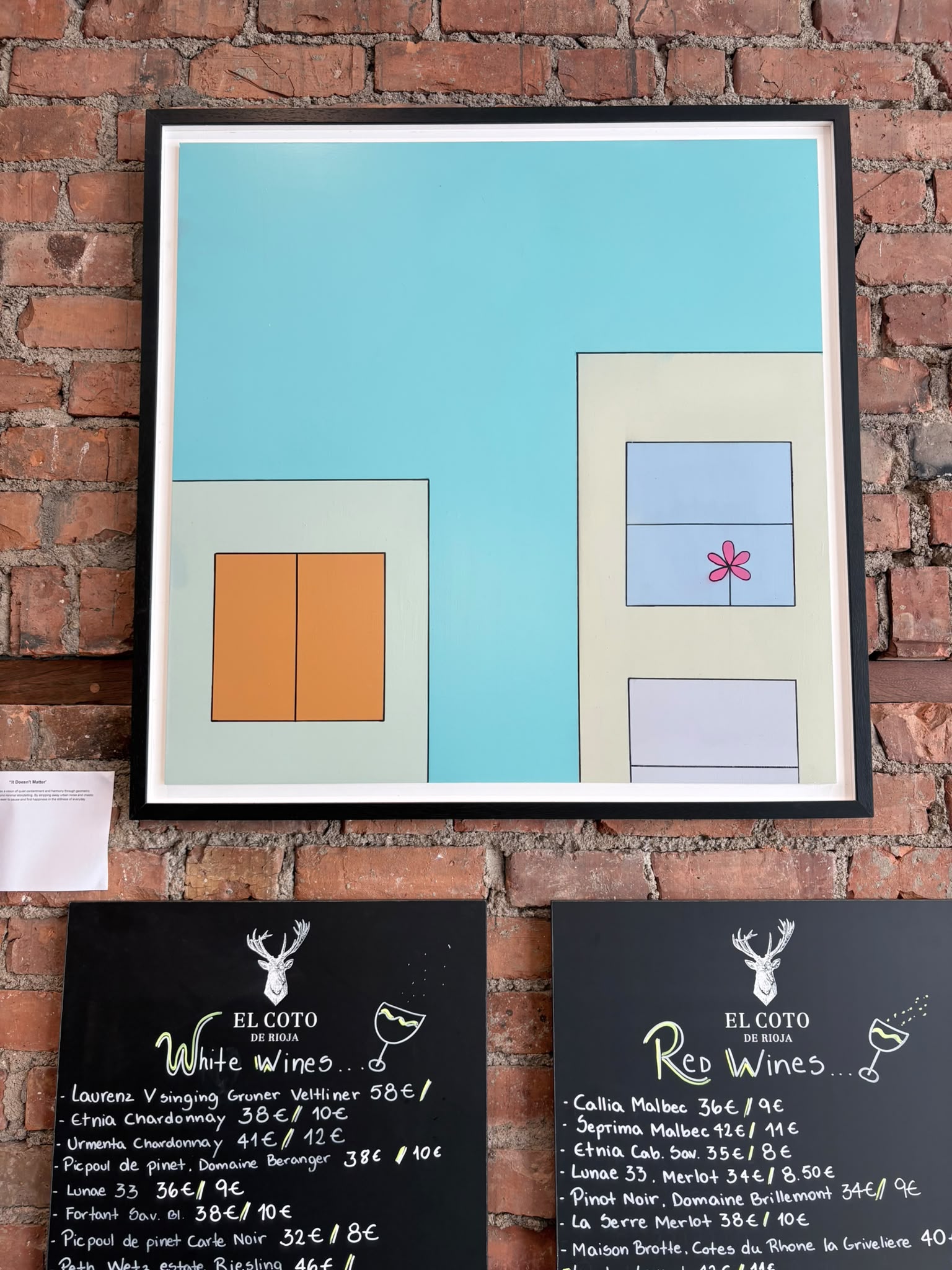 'It Doesn't Matter' by Dominic Denis - framed painting above the wine boards: turquoise sky over two pale buildings, a pink flower in one window